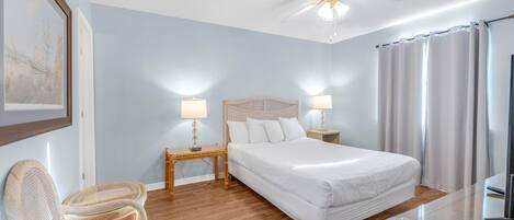 A bedroom with a bed, two bedside tables with lamps, a ceiling fan, a chair, and a glass table.