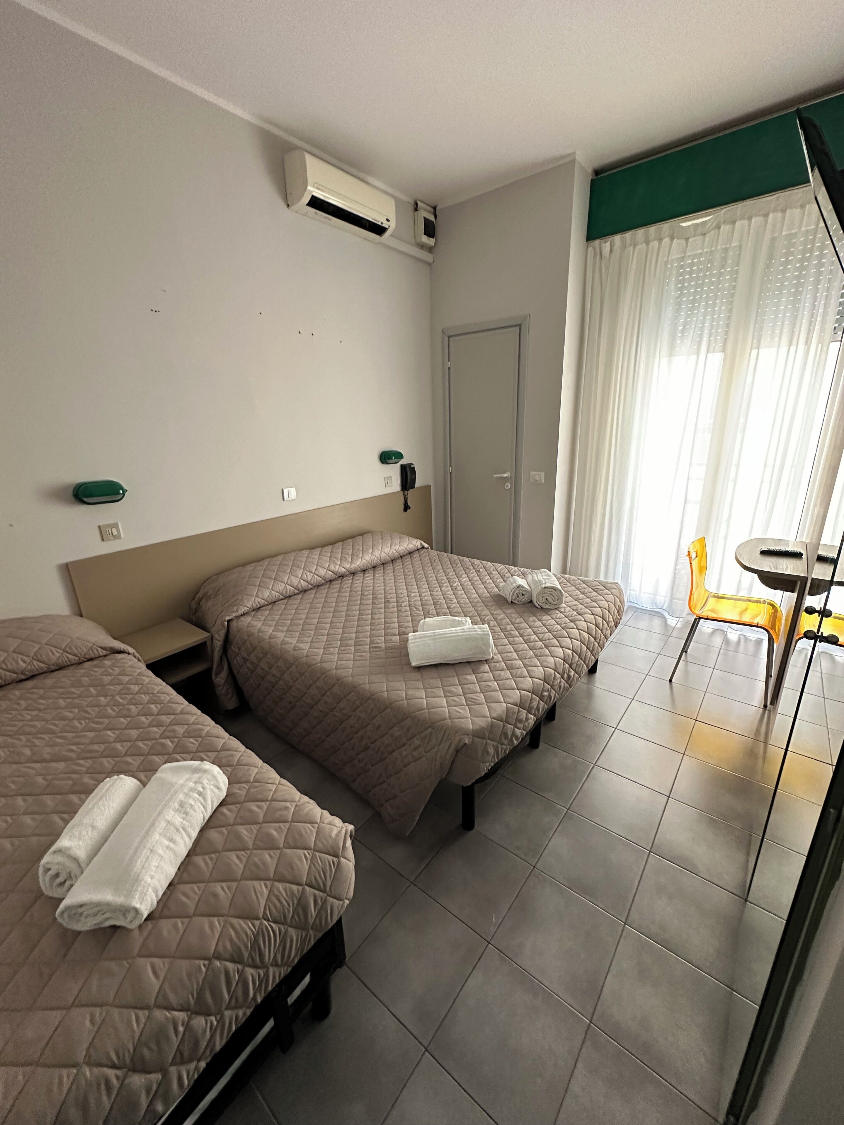 A hotel room with two beds, a small table, a chair, and a door.