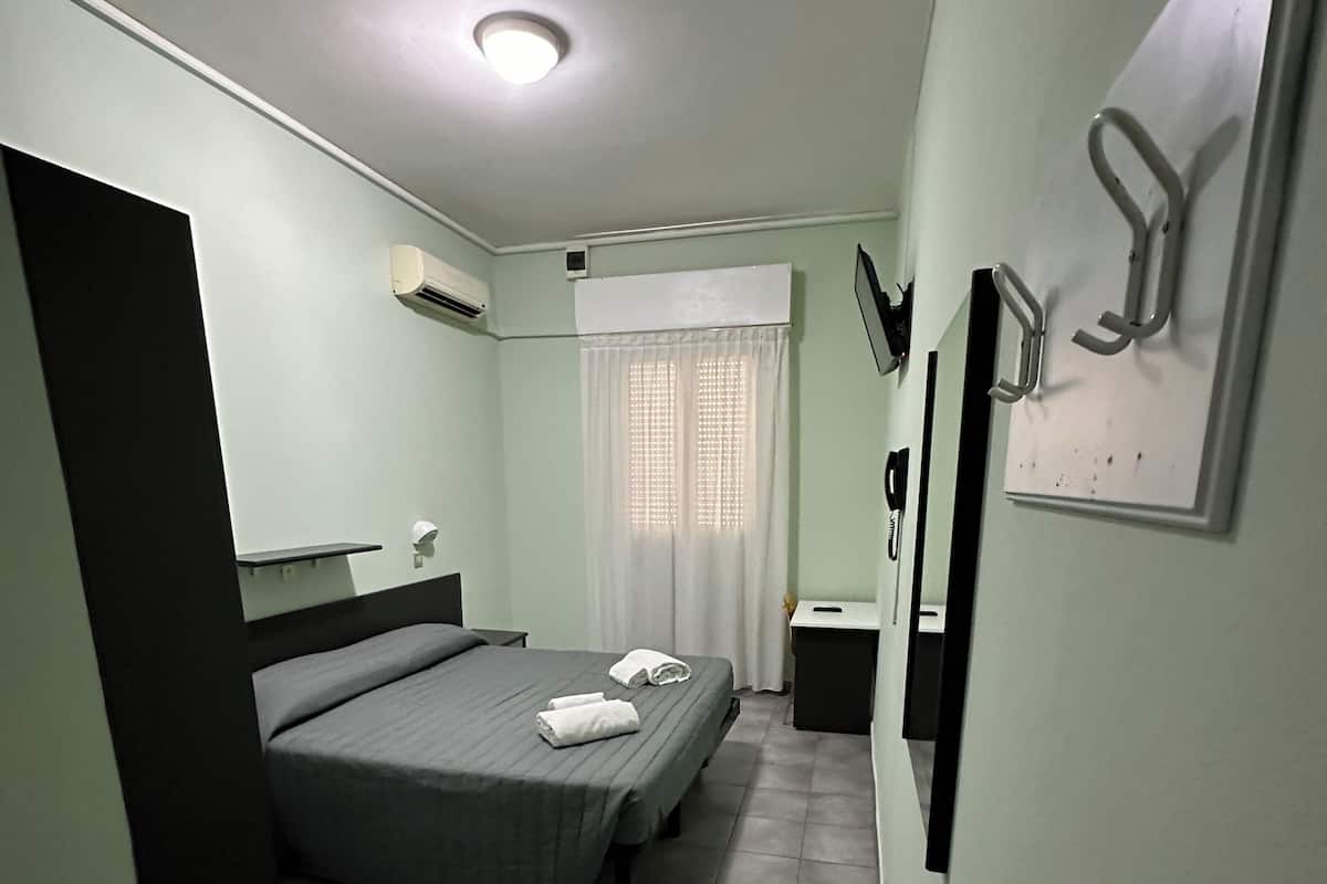 A small, well-lit bedroom with a bed, a bedside table, a mirror, and a window with curtains.