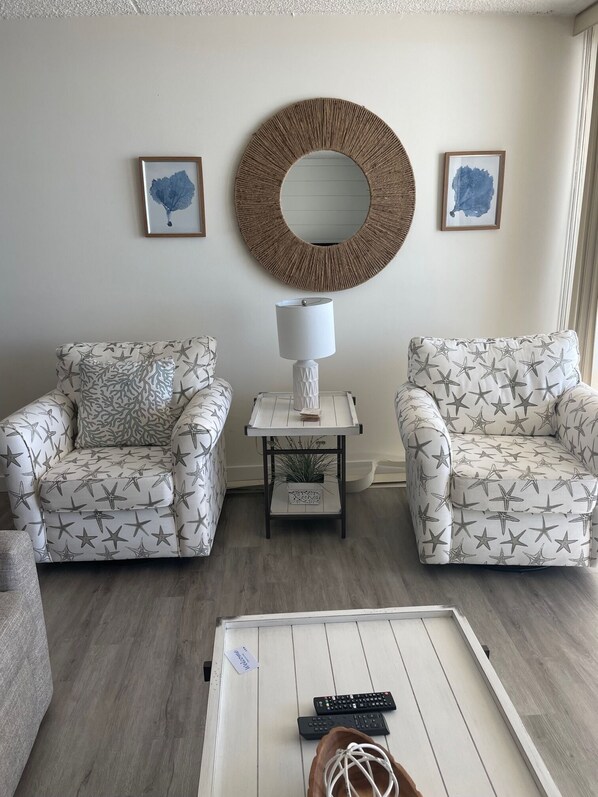 A living room with two patterned armchairs, a coffee table, a side table with a lamp, and two framed artworks on the wall.