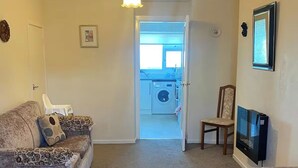 A living room with a sofa, a TV, and a chair. There is a door leading to a laundry room with a washing machine.