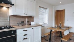 A modern kitchen with white cabinetry, a gas stove, and a wooden dining table with benches. - Cricketers View - West Yorkshire (Bradford)