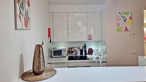 A modern kitchen with white cabinets, a microwave, toaster, and a sink. There is a decorative vase on the counter and a wall-mounted light fixture. - Liverpool Street Apartments (London)