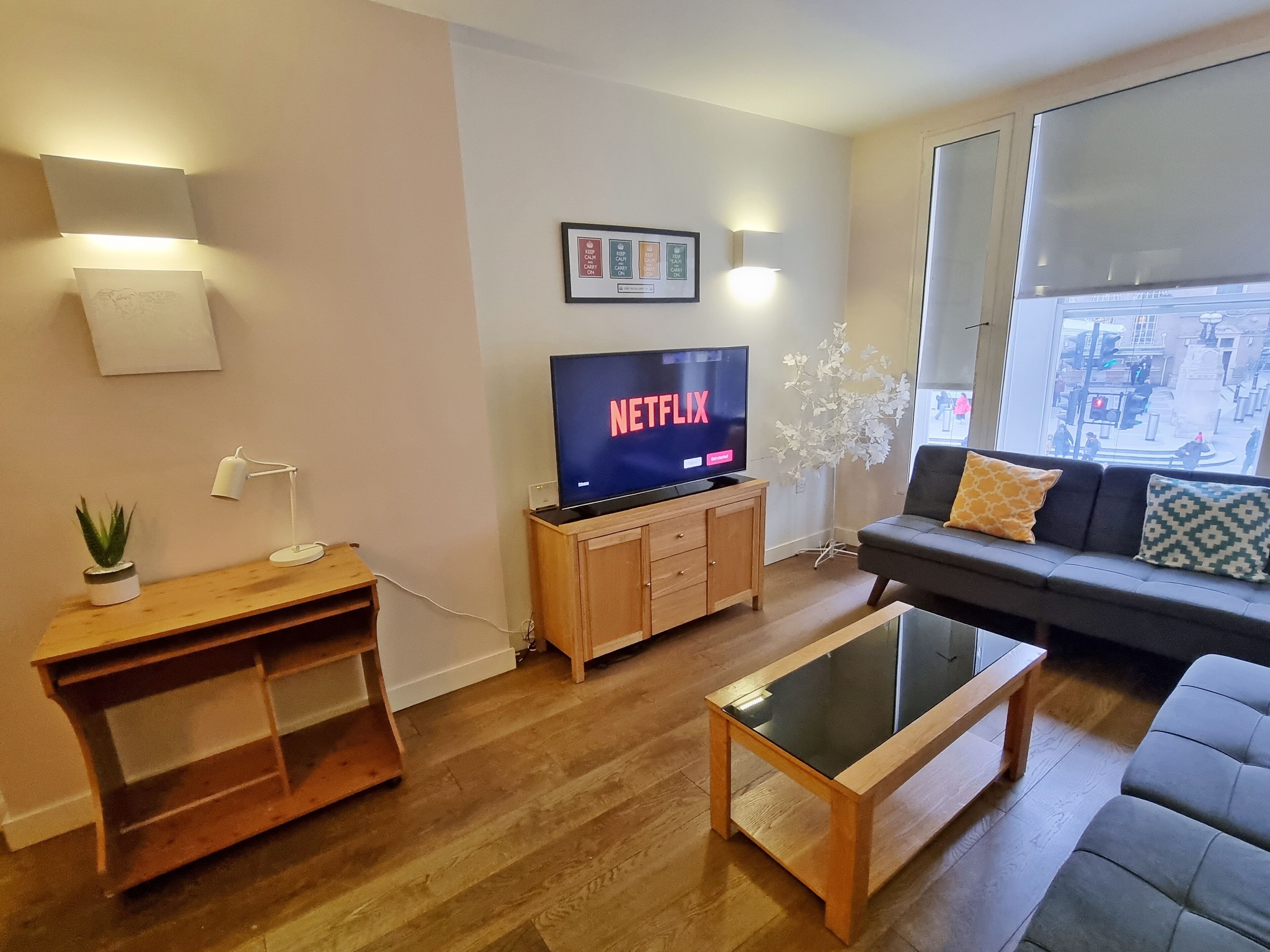 A living room with a flat-screen TV displaying Netflix, a wooden entertainment unit, a glass coffee table, a sofa with patterned cushions, and a large window offering a city view.