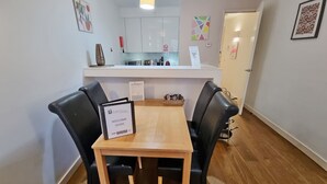 A modern kitchen with white cabinets, a microwave, and a coffee machine. There is a wooden table with two chairs in front of the counter. - Liverpool Street Apartments (London)