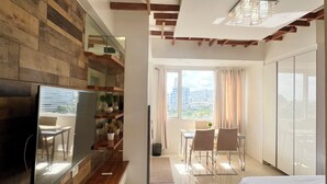 A modern hotel room with a wooden accent wall, a large bed, a dining area with a view, and a TV on the wall.