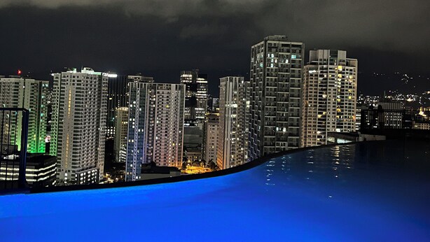 A nighttime cityscape with high-rise buildings and a pool reflecting city lights.