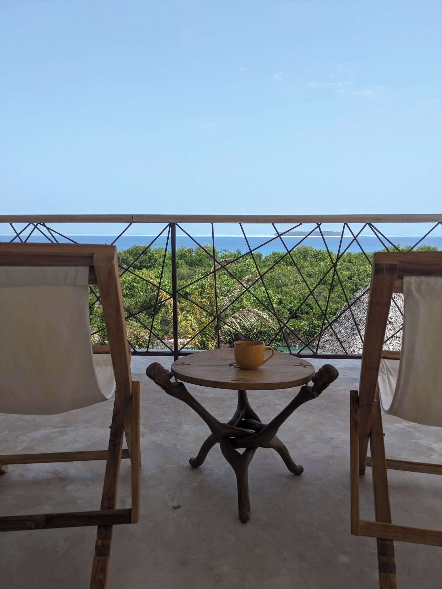 A balcony with a table and chairs, overlooking a coastal view with trees and a clear sky.