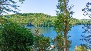Vista al lago con una terraza de madera que conduce a un muelle, rodeado de vegetación exuberante y colinas al fondo.