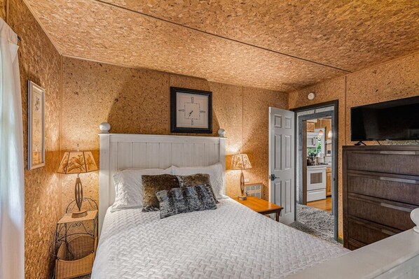 A bedroom with a wooden ceiling, a white bed with patterned pillows, bedside lamps, a clock, and a TV on a dresser.