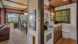 A rustic cabin interior with a dining area, kitchen, and large windows offering a view of the forest.