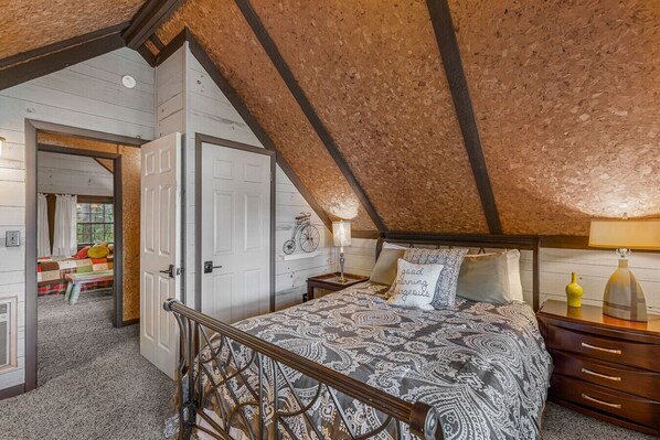 A bedroom with a sloped ceiling, a bed with patterned bedding, a nightstand with a lamp, and a door leading to another room.