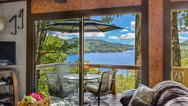 A room with a large window overlooking a lake, furnished with a table and chairs, and a TV on the left.
