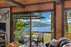 A room with a large window overlooking a lake, furnished with a table and chairs, and a TV on the left.