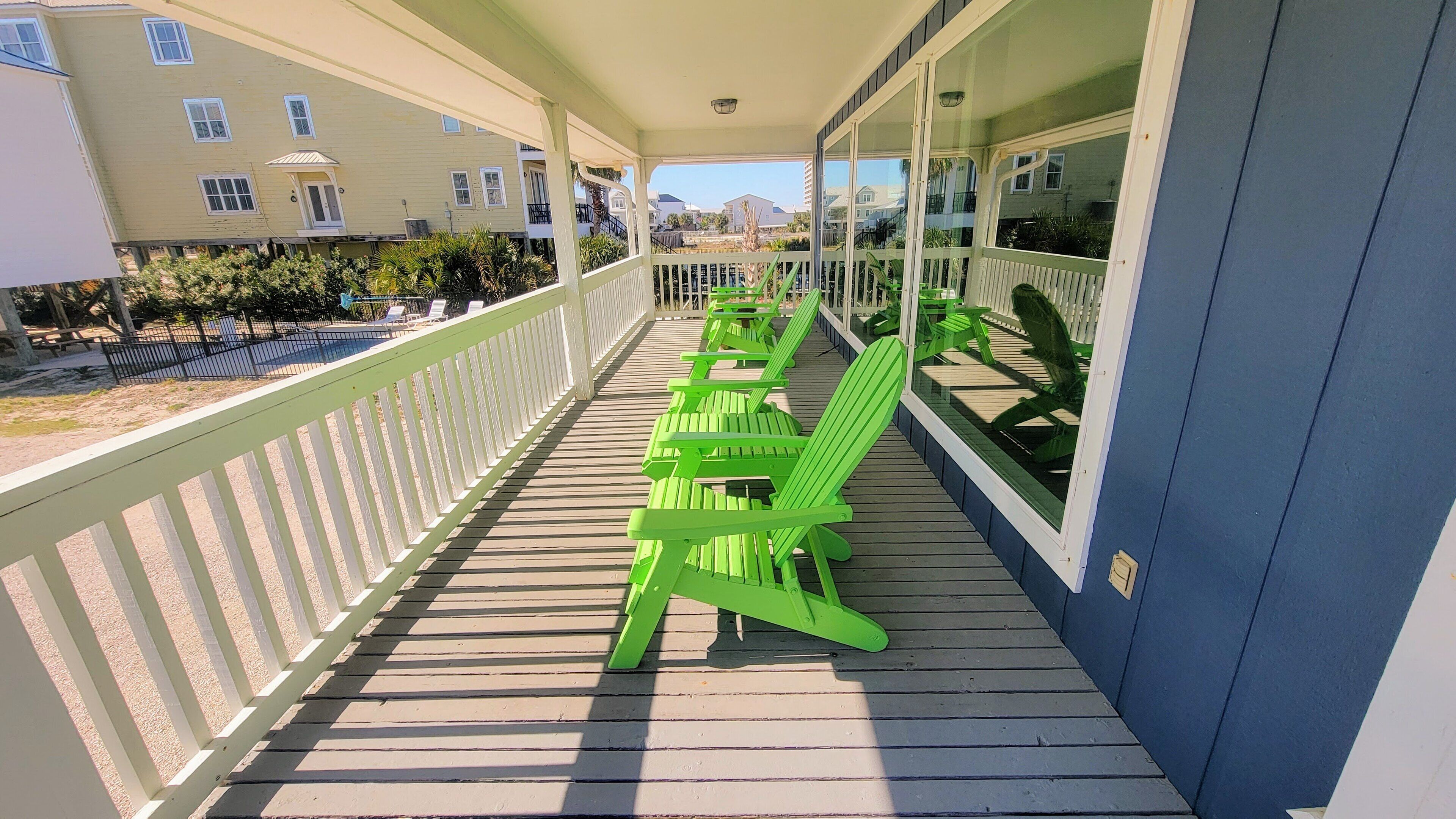 A covered outdoor area with a wooden deck, green Adirondack chairs, and a view of the surrounding buildings.