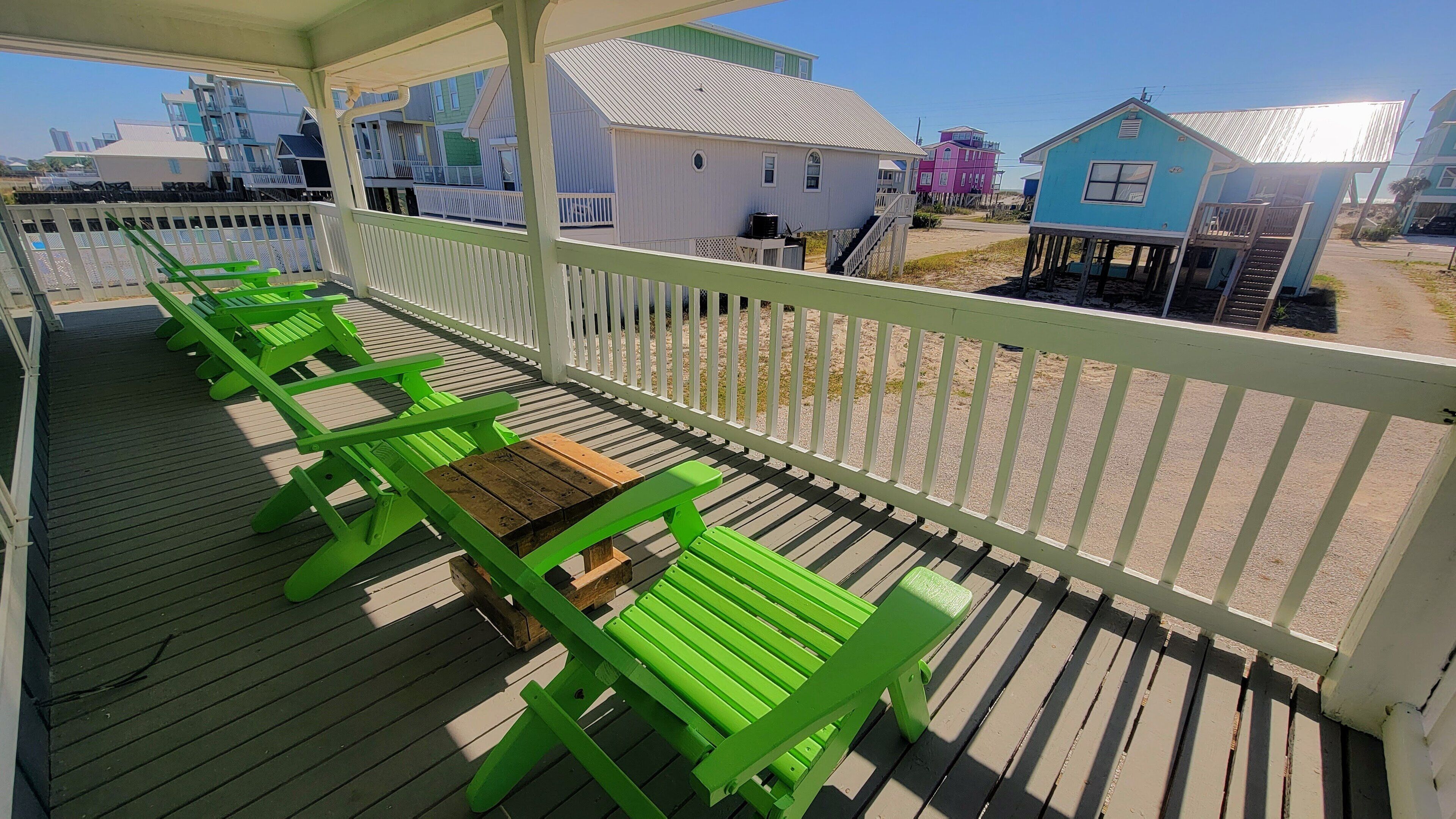 Eine überdachte Terrasse im Freien mit grünen Bänken und einem Holztisch, mit Blick auf einen Strand mit bunten Häusern.
