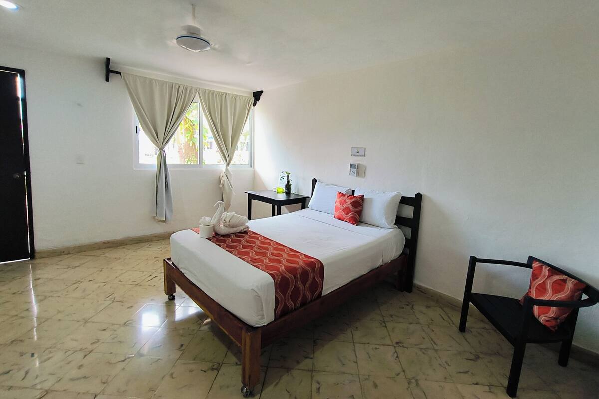A hotel room with a bed, a chair, a nightstand, a window with curtains, and a ceiling fan.