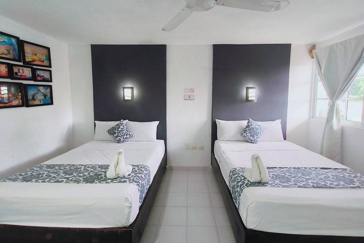 A hotel room with two beds, each with a patterned bedspread and a white pillow. There are wall-mounted lights above each bed, a ceiling fan, and a window with curtains.