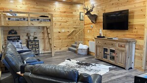 Interior acogedor de una cabaña con un sofá de cuero, una cabeza de venado montada, paredes de madera, un televisor y una escalera.
