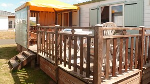 Una terraza de madera con una mesa y sillas, un espacio cubierto con un toldo amarillo, y un edificio con puerta verde.
