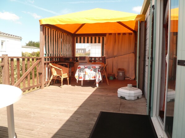 Een overdekt buitengebied met een tafel, stoelen, een kleine prullenbak en een witte koelbox.