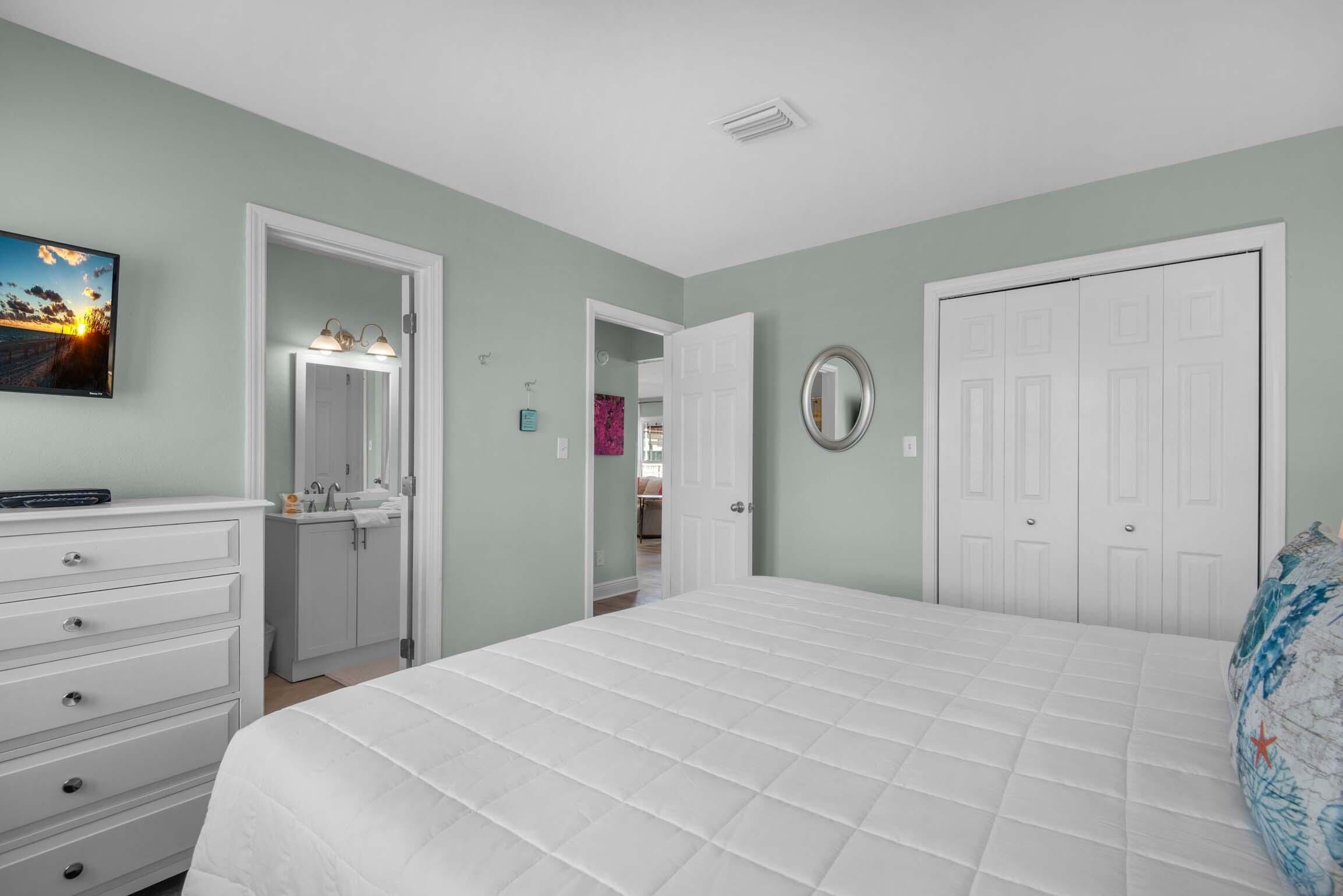 A bedroom with a large bed, a dresser, a vanity with a mirror, and a door leading to another room.