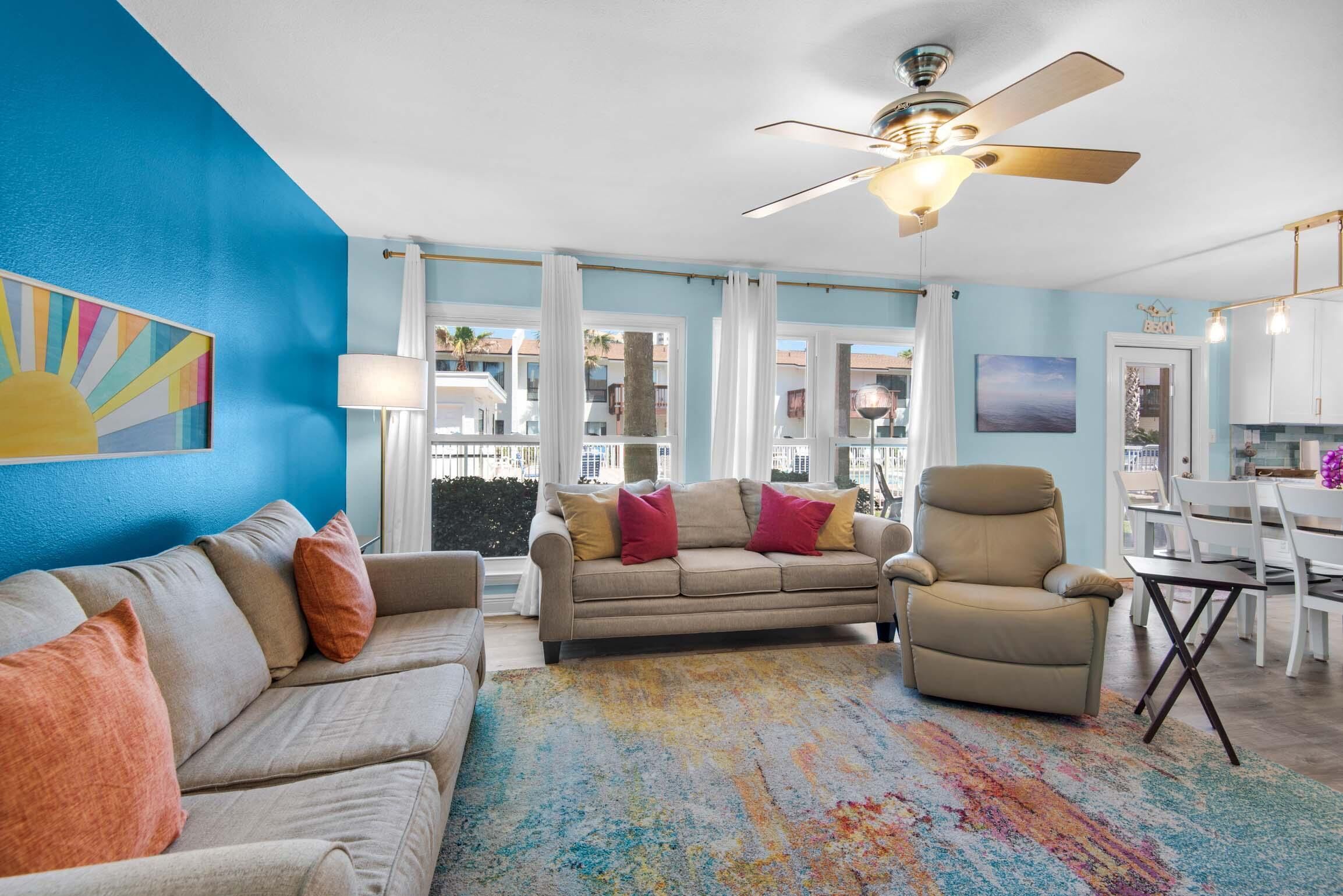 A living room with a beige sofa, a tan armchair, and a colorful rug. There is a ceiling fan, a window with curtains, and a view of buildings outside.