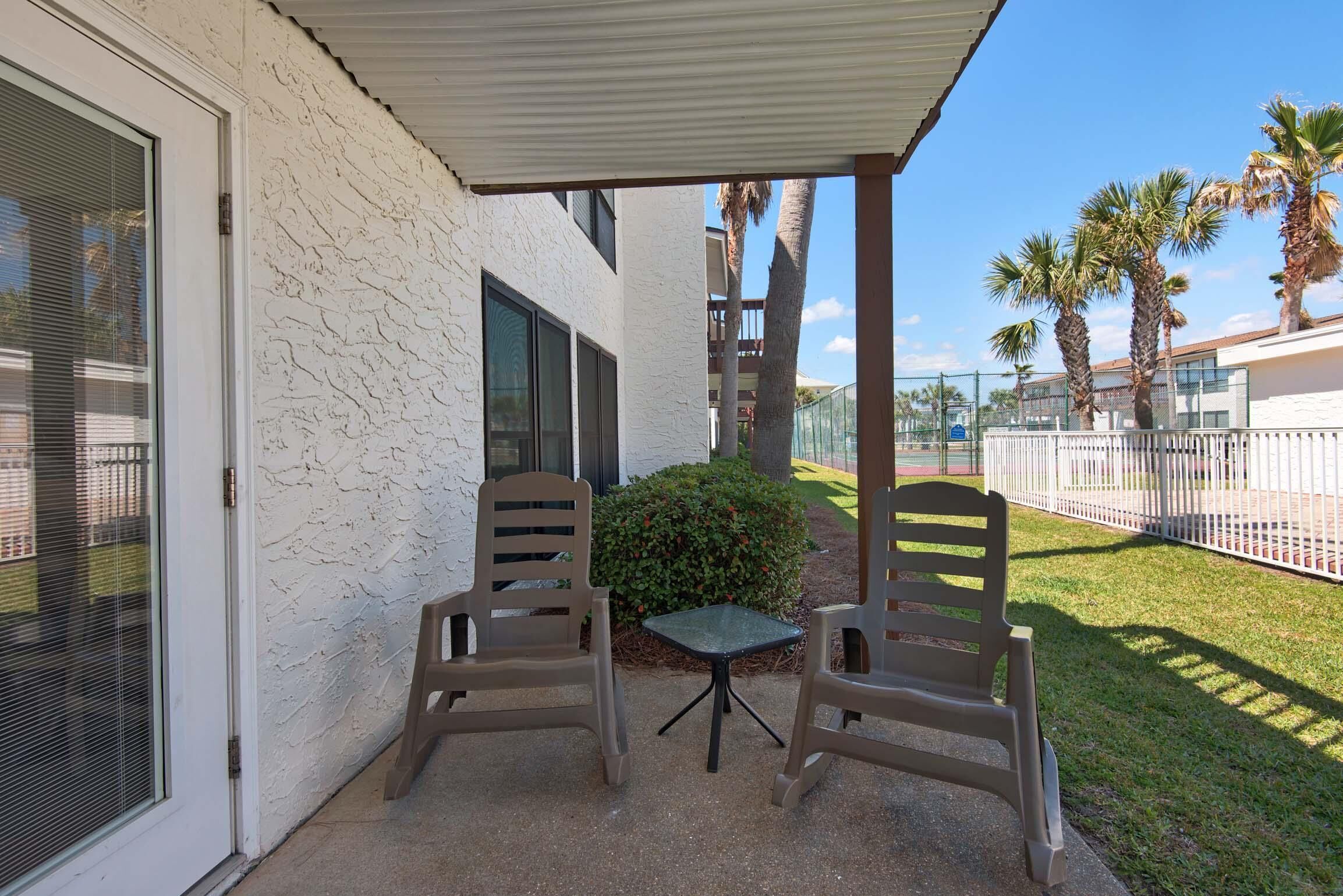 A covered patio with two chairs and a small table, a white textured wall, and a view of palm trees and a fence.
