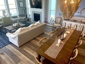A spacious living room with a large sofa, a wooden dining table with chairs, a fireplace, and a large window with a view of the beach.