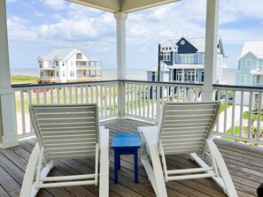 Eine überdachte Terrasse mit zwei weißen Schaukelstühlen und einem kleinen blauen Tisch, mit Blick auf einen Strand mit mehreren Häusern.