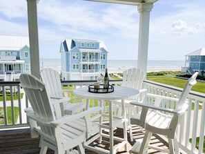 A covered outdoor seating area with white Adirondack chairs and a round table, overlooking a beach with blue houses in the background.