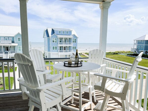 A covered outdoor seating area with white Adirondack chairs and a round table, overlooking a beach with blue houses in the background.