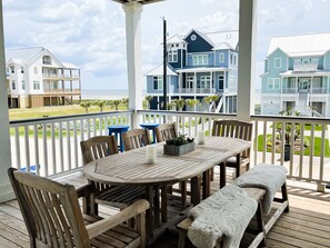 A wooden deck with a round table and benches, overlooking a beach with houses in the background.