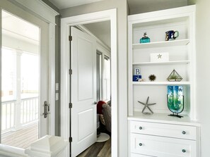 A hallway with a white door leading to a room with a wooden floor. There is a white cabinet with drawers and shelves containing decorative items.