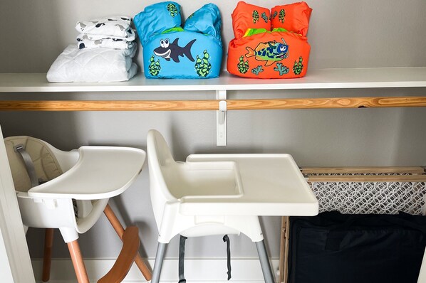 A room with two white baby high chairs, a white shelf with folded blankets, and a wooden floor.