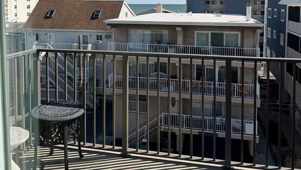 A balcony with a glass railing, a chair, and a view of adjacent buildings and the sea.