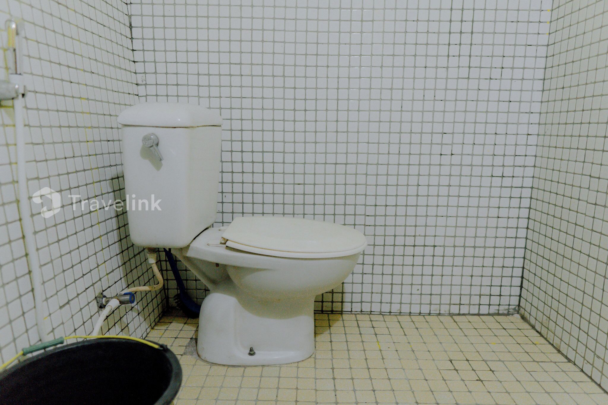 A small, tiled bathroom with a toilet, a bucket, and a pipe.