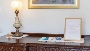 A wooden desk with a framed painting of a coastal scene, a vintage lamp, and a framed house rules sign.
