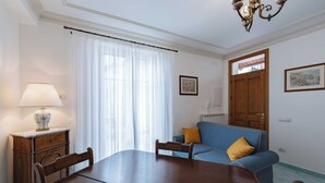 A dining area with a wooden table and chairs, a blue sofa with a yellow pillow, a lamp on a wooden side table, and a window with sheer curtains.