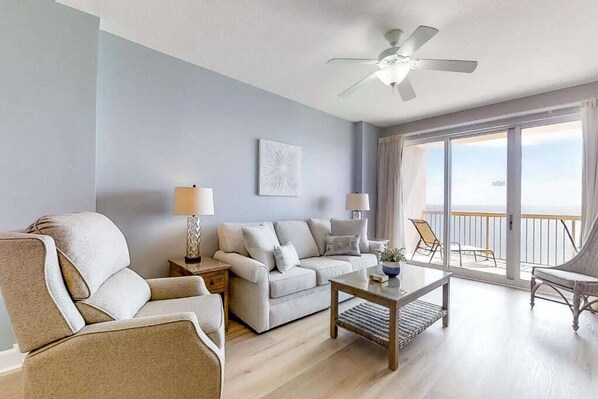 Een woonkamer met een bank, fauteuil, salontafel en een plafondventilator. Er is een balkon met uitzicht op de oceaan.