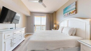 Une chambre d’hôtel avec un grand lit, un ventilateur de plafond, un téléviseur posé sur une commode, un balcon avec des rideaux et un tableau accroché au mur.