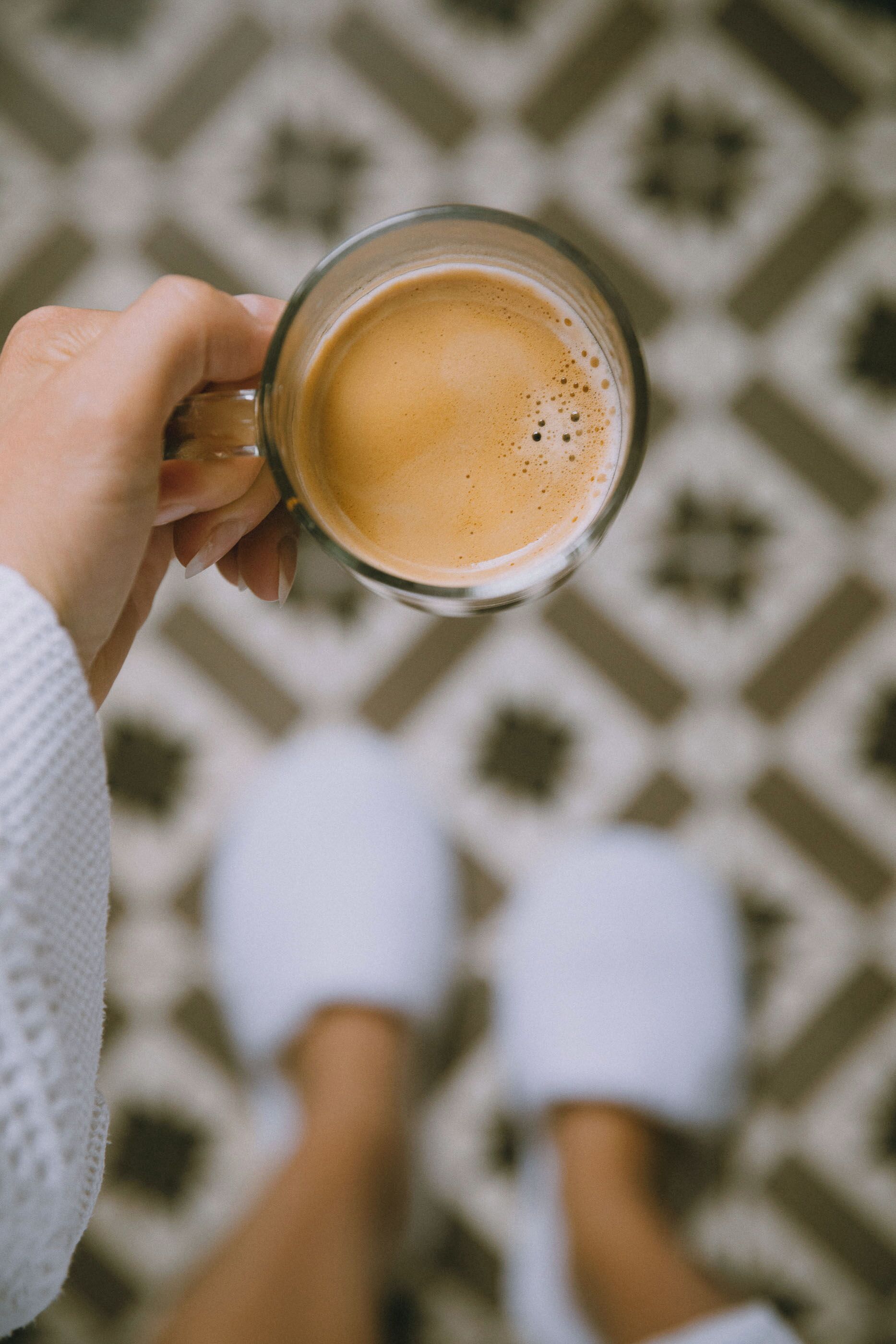 A person holding a glass mug of coffee, wearing white slippers, with a patterned floor in the background.