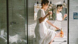 Une personne vêtue d’une robe blanche se tient dans une salle de bain, devant un miroir, un lavabo et une bouteille de champagne posée sur le comptoir.