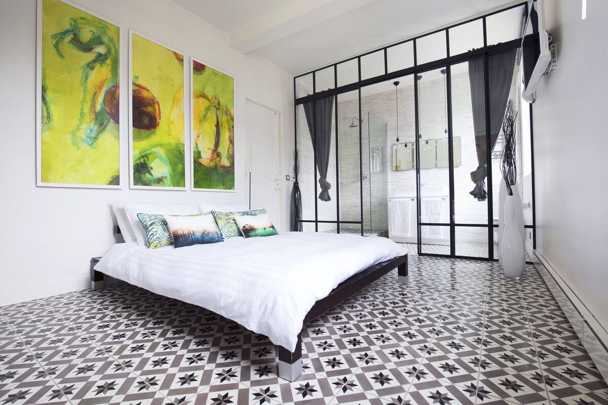 A bedroom with a bed, a large window with curtains, and abstract paintings on the wall.