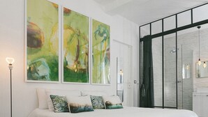 Une chambre avec un grand lit, une porte de douche en verre et des tableaux abstraits accrochés au mur.