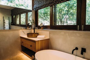 A modern bathroom with a freestanding bathtub, a round gold sink, wooden cabinetry, and large windows offering a view of trees. - Phakalane Retreat (Cape Town)