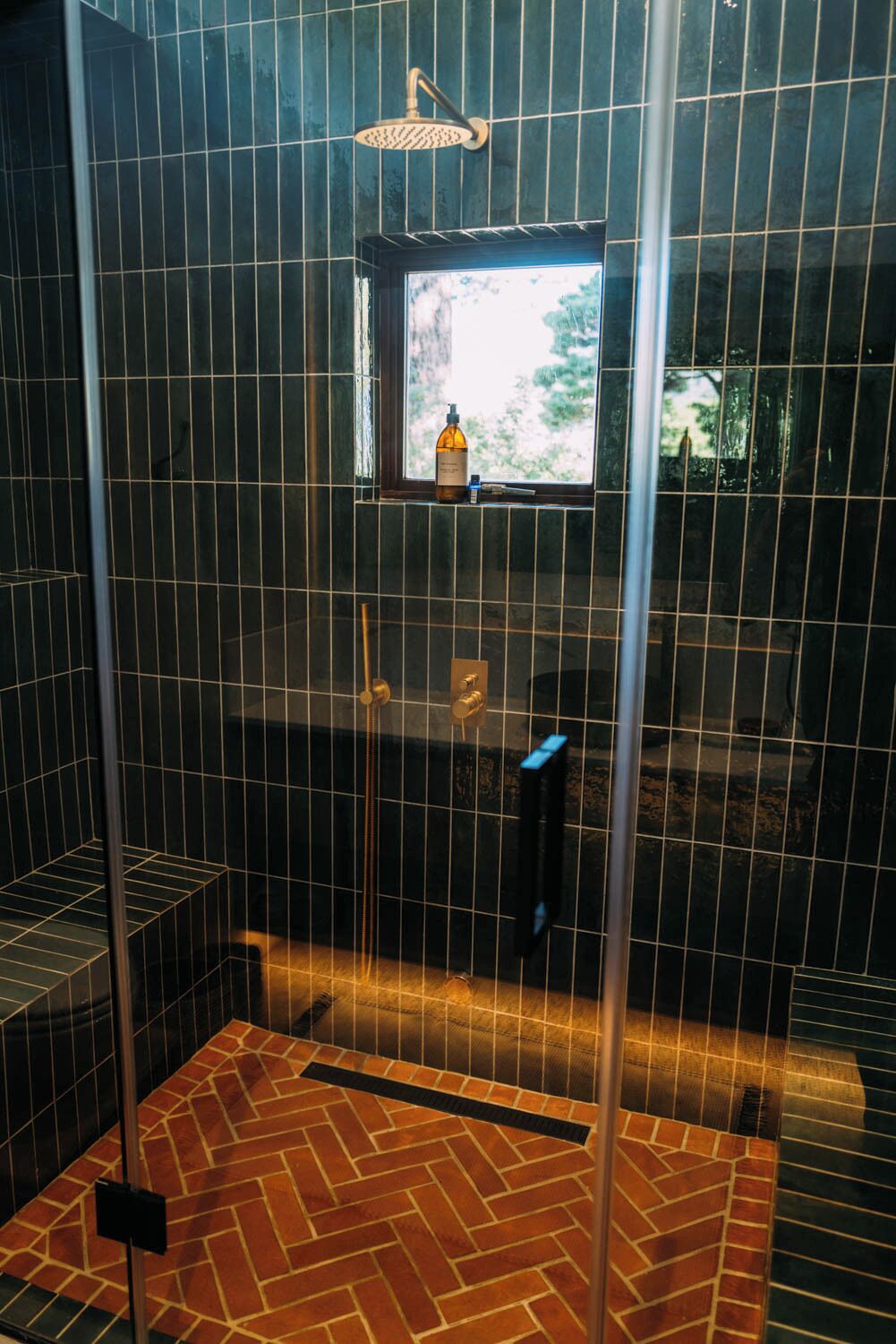 A walk-in shower with a window, a bottle of shampoo, and a handheld showerhead.