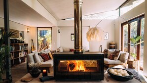 A cozy living room with a wood-burning stove, comfortable seating, a bookshelf, and a large window with a view of greenery. - Phakalane Retreat (Cape Town)