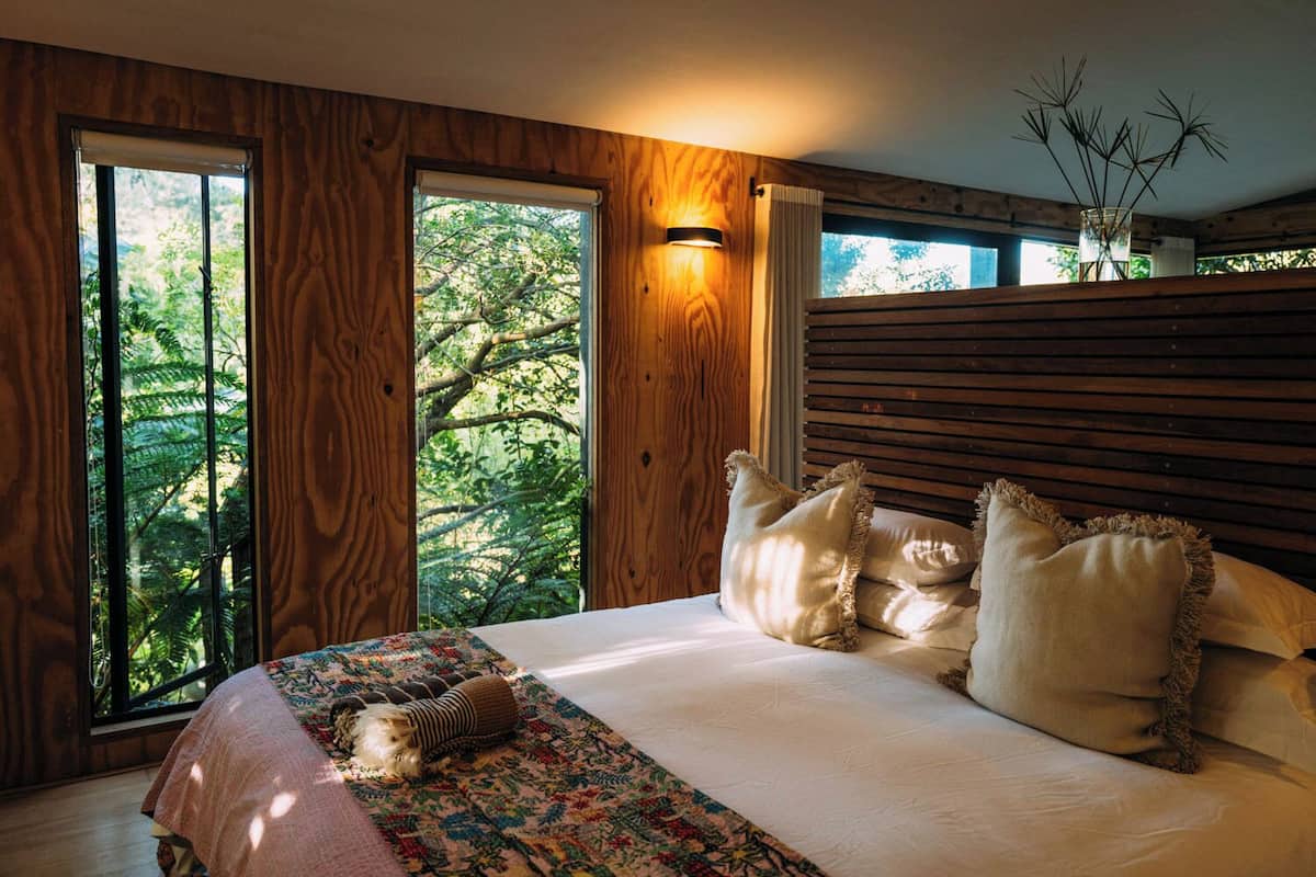 A bedroom with a wooden headboard, a bed with white linens, and a view of trees through large windows.