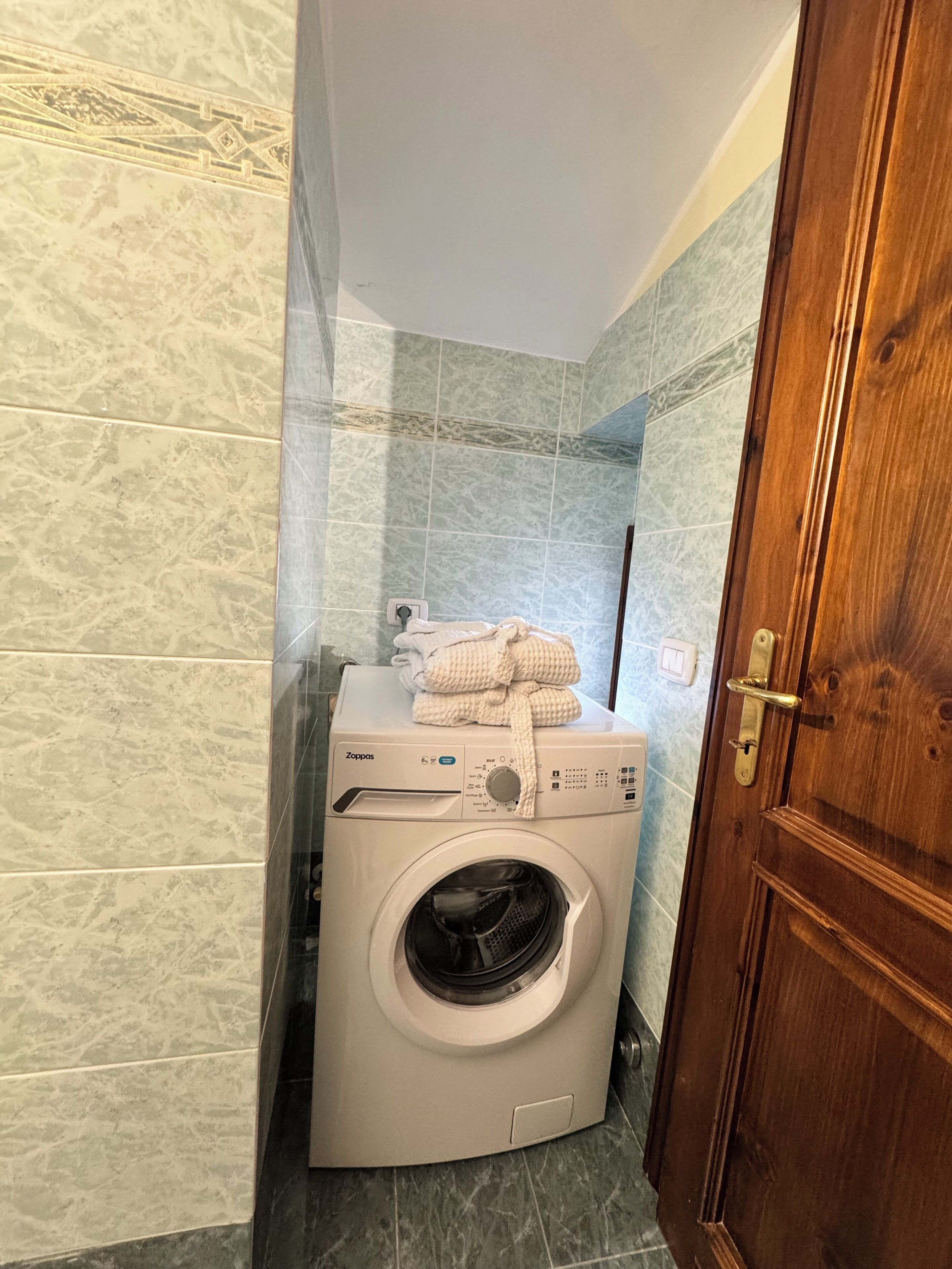 Een compacte, witte wasmachine voor eigen gebruik met een bedieningspaneel, een handdoek erboven en een houten deur aan de rechterkant.